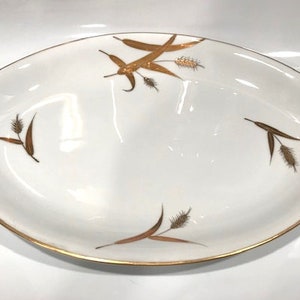 May include: White oval serving dish with gold trim and wheat stalk designs. The dish has a scalloped edge and gold accents on the handles. A decorative piece for serving food or displaying as a decorative item.