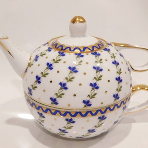 Op de afbeelding: Een witte porseleinen theepot en kop en schotel set. De set heeft een blauw bloemmotief met gouden accenten. De theepot is rond met een tuit en handvat. De kop past eronder.