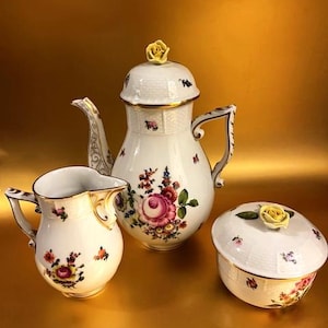 May include: A white porcelain tea set with floral patterns. The set includes a teapot, creamer jug, and a covered sugar bowl. The teapot has a yellow rose on the lid. The creamer and sugar bowl also feature floral patterns and gold trim.