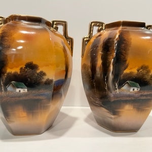 May include: Two matching decorative vases with a landscape scene. The vases have a gold trim and handles. The landscape features a small white house with a green roof, trees, and a body of water, all set against a warm orange sky.
