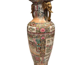 May include: A tall, ornate porcelain vase with a wide mouth and a bulbous body. The vase features a detailed, hand-painted design with floral and figural scenes in shades of pink, green, and gold. Gold elephant head handles adorn the sides.