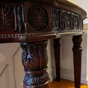 May include: A dark brown, antique wooden table with intricate carvings. The table features a marble top and ornate detailing along the apron and legs. The legs are fluted and carved with floral motifs, showcasing a classic, elegant design.