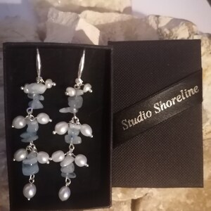 Puede incluir: Un par de pendientes colgantes con cuentas de aguamarina y perlas. Los pendientes están en una caja negra con el texto "Studio Shoreline" en la parte superior.