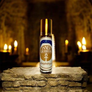 May include: A roll-on bottle with a gold cap and a label featuring the text "Inner Goddess Apothecary" and the name "Nephthys". The label has a blue and white star design and an ankh symbol. The bottle is on a stone surface.