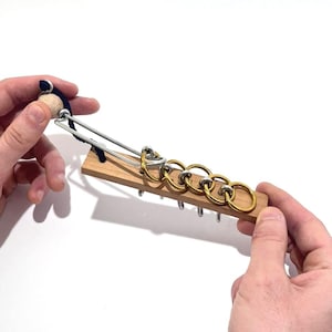 Può includere: Un puzzle in legno con anelli di metallo e una piccola maniglia in legno. Il puzzle presenta anelli ad incastro attaccati a una base di legno. Gli anelli sono in varie tonalità di oro e argento. La maniglia è bianca e nera.