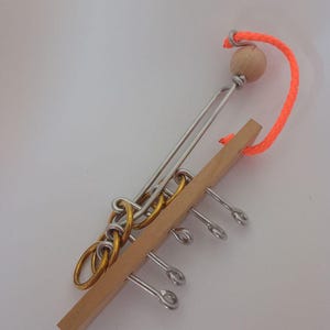Può includere: Un puzzle in legno e metallo con base in legno, anelli in metallo e un cordino arancione brillante. Il puzzle presenta pezzi metallici ad incastro e una sfera di legno. Il design è un rompicapo.
