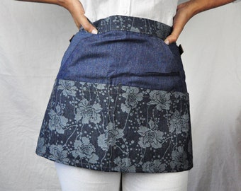 May include: A denim half-apron with a floral pattern. The apron is a dark blue color with a lighter blue floral design on the bottom. The apron is worn over white pants and a white button-down shirt.