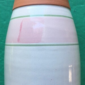 May include: A handmade ceramic vase with a light blue base, a pink band, and a terracotta rim. The vase features green stripes and a unique, rounded shape. The bottom has a stamped design.