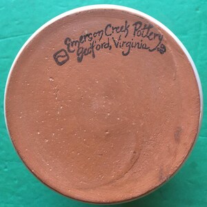 May include: The bottom of a ceramic mug with a white rim and a reddish-brown base. The text "Emerson Creek Pottery Bedford, Virginia" is handwritten in black ink on the base of the mug.
