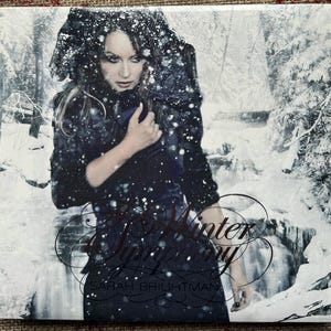 May include: Album cover featuring Sarah Brightman in a dark, winter-themed setting. She is wearing a dark outfit with a snowy effect overlay. The title "Winter Symphony" is written in an elegant script, with the artist's name below.