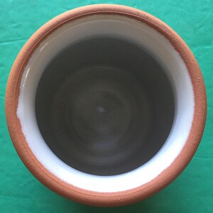 May include: A handmade ceramic vessel with a wide opening. The rim is a warm terracotta color, with a white band on the inside edge. The interior is a dark gray, creating a contrast with the lighter rim. The vessel is likely used for holding liquids or as a decorative item.