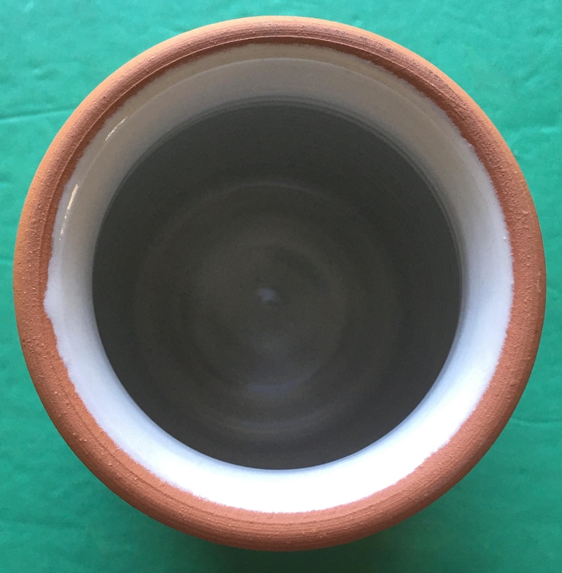 May include: A handmade ceramic vessel with a wide opening. The rim is a warm terracotta color, with a white band on the inside edge. The interior is a dark gray, creating a contrast with the lighter rim. The vessel is likely used for holding liquids or as a decorative item.