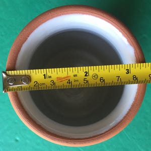 May include: A handmade ceramic pot with a brown exterior, white interior rim, and gray interior. The pot's diameter is approximately 3 inches. A yellow measuring tape is placed across the top of the pot.