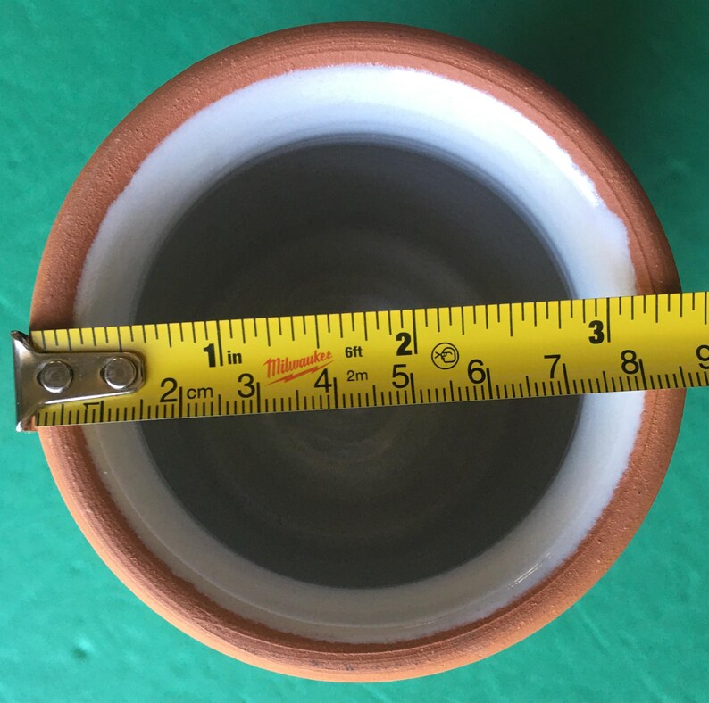 May include: A handmade ceramic pot with a brown exterior, white interior rim, and gray interior. The pot's diameter is approximately 3 inches. A yellow measuring tape is placed across the top of the pot.