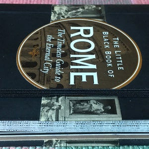 Pu&ograve; includere: Un libro nero con un adesivo dorato che dice "The Little Black Book of Rome" e "The Timeless Guide to the Eternal City". Il libro ha un elastico nero e un adesivo verde che dice "Includes fold out maps".