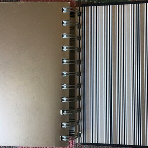 Pu&ograve; includere: Un quaderno color oro con una copertina a righe bianche e nere. Il quaderno ha una rilegatura a spirale in metallo.