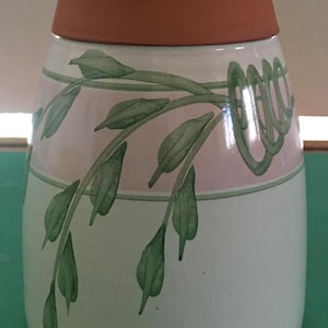 May include: A ceramic vase with a wide, terracotta-colored rim and a white body. The vase is decorated with a pink and green band featuring a stylized leaf and vine design. The leaves are green, and the vase has a rounded shape.