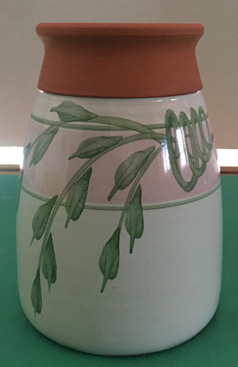 May include: A ceramic vase with a wide, terracotta-colored rim and a white body. The vase is decorated with a pink and green band featuring a stylized leaf and vine design. The leaves are green, and the vase has a rounded shape.