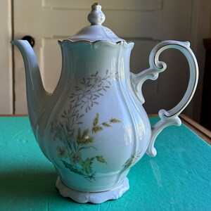 May include: A white porcelain coffee pot with a lid, spout, and handle. The pot is decorated with gold trim and floral designs in green and yellow. The pot has a height of approximately 9 inches.