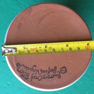 May include: A round, brown pottery piece with a white rim. A yellow tape measure shows a diameter of approximately 10 inches. The text "Emerson Creek Pottery, Bedford Virginia" is written on the pottery.
