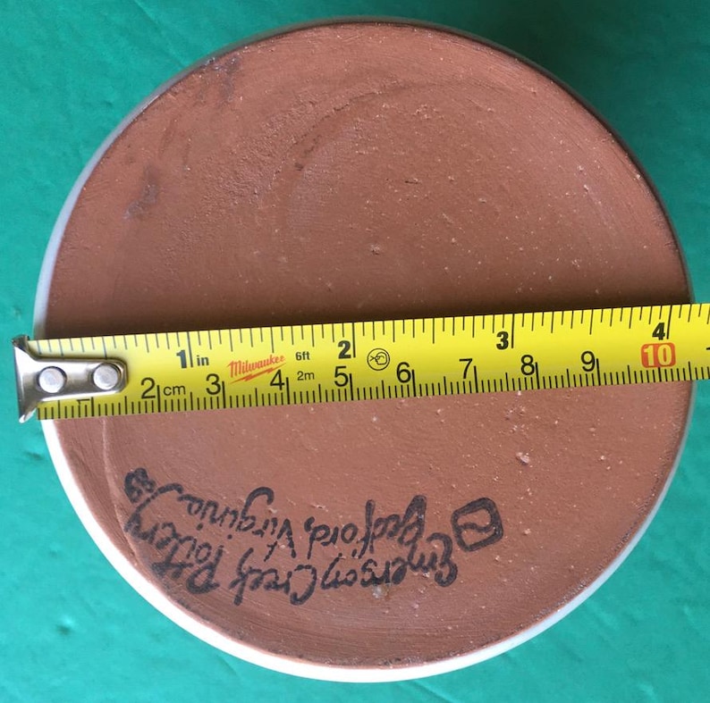May include: A round, brown pottery piece with a white rim. A yellow tape measure shows a diameter of approximately 10 inches. The text "Emerson Creek Pottery, Bedford Virginia" is written on the pottery.