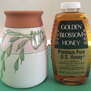 May include: A ceramic jar with a terracotta rim and a white body decorated with pink bands and green leaf designs. Next to it is a bottle of Golden Blossom Honey, containing 24 oz (1 lb 8 oz) of honey.