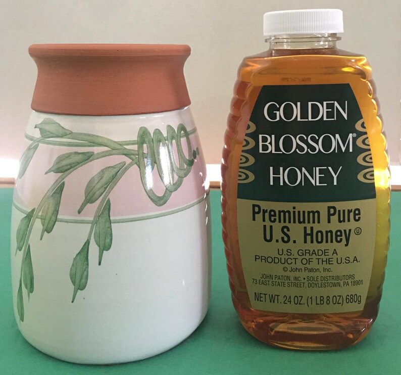 May include: A ceramic jar with a terracotta rim and a white body decorated with pink bands and green leaf designs. Next to it is a bottle of Golden Blossom Honey, containing 24 oz (1 lb 8 oz) of honey.