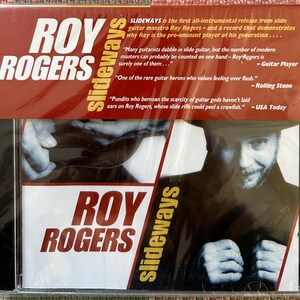 May include: A CD case for Roy Rogers' album "SlideWays." The album cover features the artist's name in large white letters and the album title in yellow. The image includes quotes from "Guitar Player," "Rolling Stone," and "USA Today."
