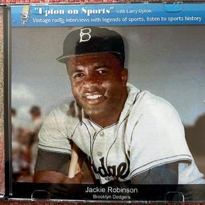May include: CD case featuring Jackie Robinson of the Brooklyn Dodgers. The image shows Robinson in his baseball uniform, smiling. The CD case has the title "Upton on Sports" with text about vintage radio interviews.