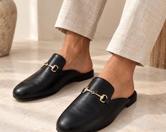 May include: A pair of black leather backless loafers featuring a gold-toned metal horsebit detail across the vamp. The shoes have a rounded toe, a low heel, and a smooth finish, worn with light-colored trousers that end above the ankle.