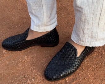 May include: A pair of black woven leather loafers with a slip-on design and a low heel, paired with white textured trousers. The shoes feature a detailed basket-weave pattern across the upper surface and a smooth, dark sole.