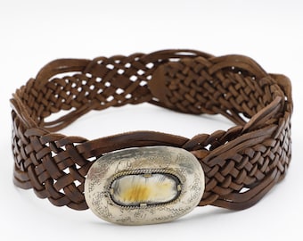 May include: A brown leather belt with a woven design. The belt features a silver-colored oval buckle with a light-colored stone inlay. The belt is displayed on a white background.