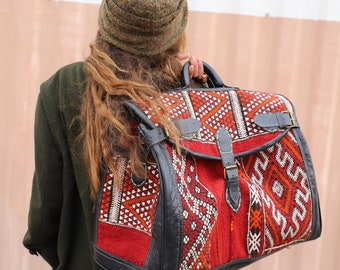 May include: A woman wearing a green hat carries a large, red, black, and white patterned leather bag with a buckle closure.