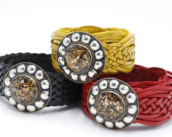 May include: Three braided leather bracelets in black, yellow, and red. Each bracelet features a large, round, silver-toned buckle with a central, textured, amber-colored stone. The bracelets are displayed against a white background.