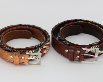 May include: Two belts with brown leather and colorful woven fabric. The belts have silver-colored buckles. The woven fabric features a geometric pattern with red, green, and white accents. The belts are coiled on a white background.