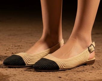 May include: A pair of beige and black slingback flats. The shoes have a woven texture on the upper and a black toe cap. A small gold buckle secures the slingback strap. The shoes are on a dirt surface.