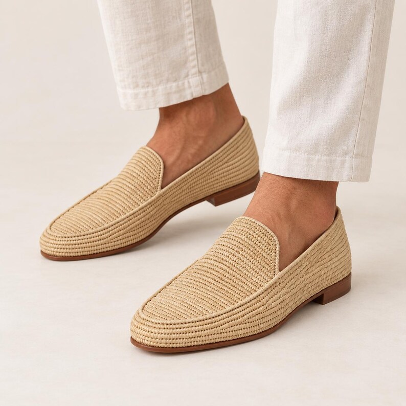 May include: A pair of beige woven loafers featuring a textured straw-like material, a rounded toe, and a low brown heel. The slip-on shoes have a structured silhouette with a clean finish, paired with white trousers that end above the ankle.