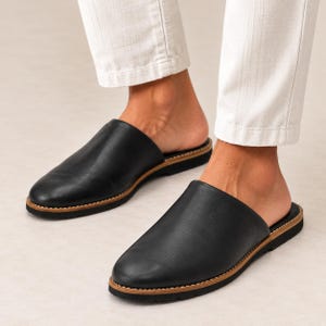 May include: A pair of black leather slip-on mules featuring a rounded toe, a tan midsole with visible white stitching, and a black textured outsole. The footwear has an open-back design and a smooth, matte finish across the upper surface.