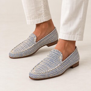 May include: A pair of slip-on loafers featuring a woven texture in alternating blue and cream threads. The shoes have a low-profile silhouette with a rounded toe, a visible stitched seam along the top, and a flat brown sole with a small heel.