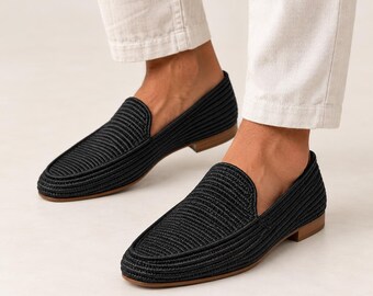 May include: A pair of black loafers featuring a textured, woven cord upper design with a rounded toe, a low heel, and a flat tan sole. The shoes are worn with light-colored trousers, showcasing the slip-on style and the detailed ribbed pattern.
