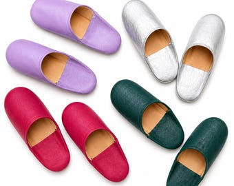 May include: A collection of eight slip-on slippers arranged in pairs, featuring smooth leather-like uppers in lavender, metallic silver, deep magenta, and dark forest green, all with tan-colored interior linings and rounded toe designs.