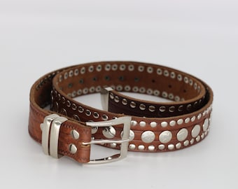 May include: Brown leather belt with silver-tone studs and a silver buckle. The belt features a series of small, evenly spaced studs along the edges and larger, decorative studs in the center. A classic accessory.