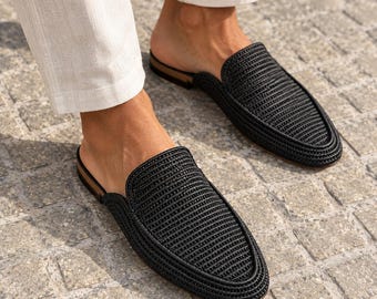 May include: A pair of black woven slip-on mules featuring a textured, corded upper design with a rounded toe and a low-profile silhouette. The shoes have a dark brown heel base and are worn with light-colored trousers, showcasing a slip-on style.