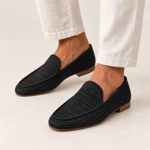 May include: A pair of black loafers featuring a textured, woven cord upper design with a rounded toe, a low heel, and a flat tan sole. The shoes are worn with light-colored trousers, showcasing the slip-on style and the detailed ribbed pattern.