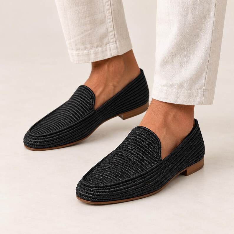 May include: A pair of black loafers featuring a textured, woven cord upper design with a rounded toe, a low heel, and a flat tan sole. The shoes are worn with light-colored trousers, showcasing the slip-on style and the detailed ribbed pattern.