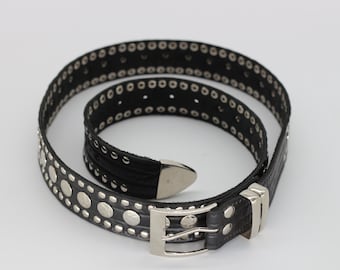 May include: Black leather belt with silver-colored studs and eyelets. The belt has a silver buckle and a pointed end. The belt is coiled on a white surface.