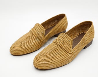 May include: A pair of tan woven loafers featuring a textured straw-like exterior, a classic slip-on silhouette with a rounded toe, and a low brown heel. The interior lining is dark brown, and the edges show a decorative serrated trim detail.