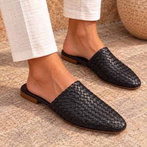 Peut inclure: Une paire de mules en cuir tressé noir avec un bout pointu et une semelle plate marron. Ces chaussures à enfiler présentent une texture en tressage panier détaillée sur la tige, offrant un aspect structuré avec un design plat.
