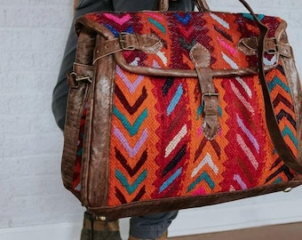 May include: A large, colorful, woven bag with leather accents and a buckle closure. The bag has a geometric pattern in shades of red, orange, pink, blue, and green. The bag is being held by a person wearing a dark jumpsuit.