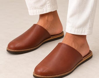 May include: A pair of brown leather slip-on mules featuring a rounded toe, a smooth upper surface, and a dark brown sole with visible white stitching along the welt. The footwear is worn with white trousers, showcasing a backless design.
